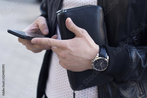 young man wearing leather jacket and wrist watches holding a leather wallet and using a smartphone 