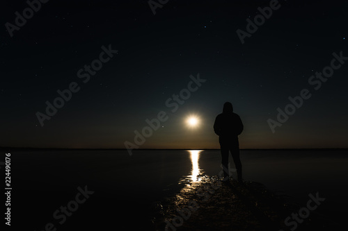 The man stands at the sunset of the moon under the starry sky with bright stars and milk way. Silhouetted photo against the starry night sky. Reflection of the rays from the moon in the water. © Aliaksandr Marko