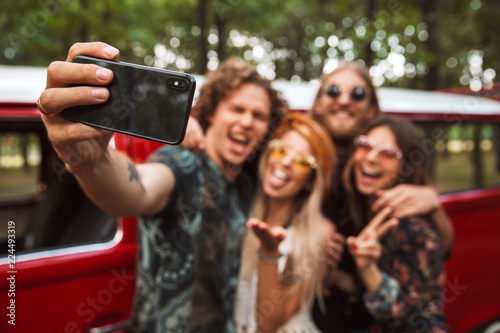Group of european hipsters men and women smiling, and taking selfie on mobile phone near vintage minivan into the nature