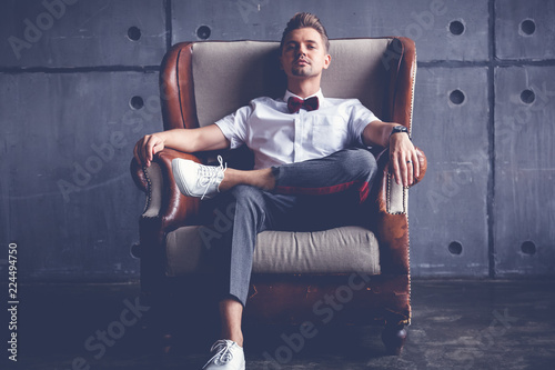 Young attractive serious glamorous male hipster in white shirt and butterfly sitting in chair, on gray background