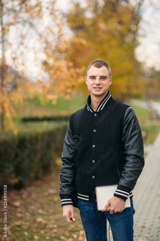 Stylish guy in black jucket stand in park. He hold a notebook in hands Stylish guy in black jucket stand in park. He hold a notebook in hands