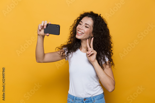 Image of european woman 20s with curly hair holding smartphone and taking selfie photo, isolated over yellow background