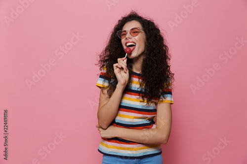 Happy cute young woman posing isolated over pink background eat candy lollipop.