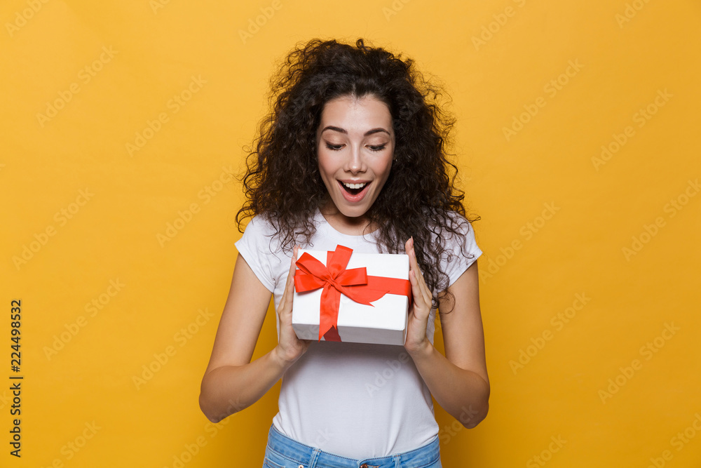 Happy cute young woman posing isolated over yellow background holding gift box present. Happy cute young woman posing isolated over yellow background holding gift box present.