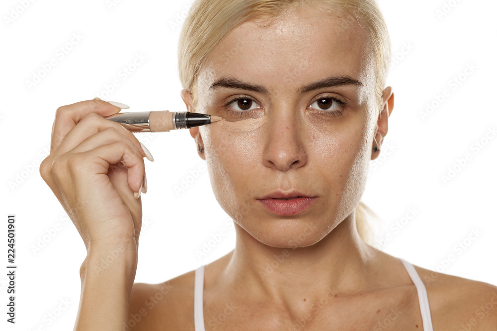 Fototapeta premium Young woman applying concealer under her eyes with brush on white background