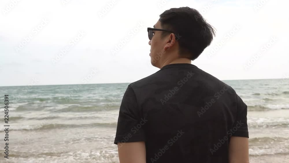Back of young man wearing sunglasses, standing and looking at the blue sea on the beach on cloudy day alone Back of young man wearing sunglasses, standing and looking at the blue sea on the beach on cloudy day alone