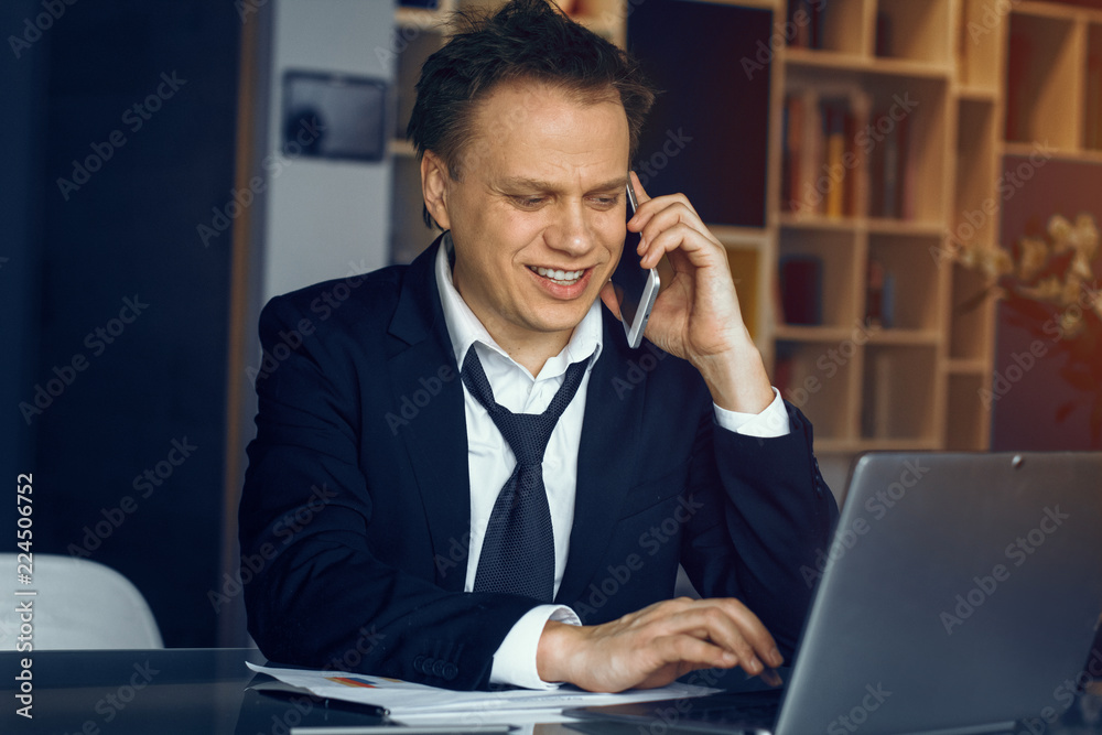 Smiling businessman sitting at table and speaking over phone. Modern laptop on table before man. Smiling businessman sitting at table and speaking over phone. Modern laptop on table before man.