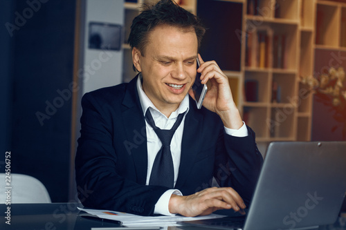 Smiling businessman sitting at table and speaking over phone. Modern laptop on table before man.