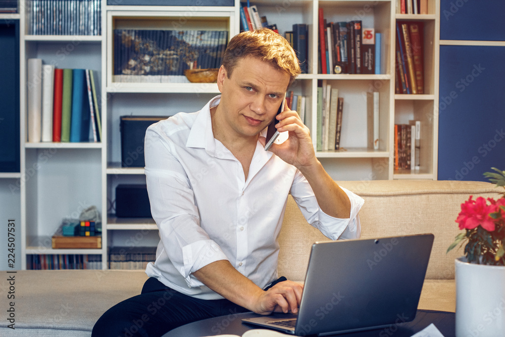 Fototapeta premium Handsome businessman in white shirt sitting in front of laptop and speaking over phone. Business concept.
