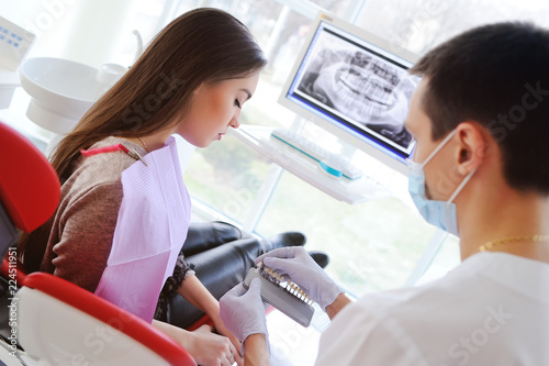 The dentist selects a young pretty girl for the patient a shade of teeth and shows on the monitor an x-ray of teeth.