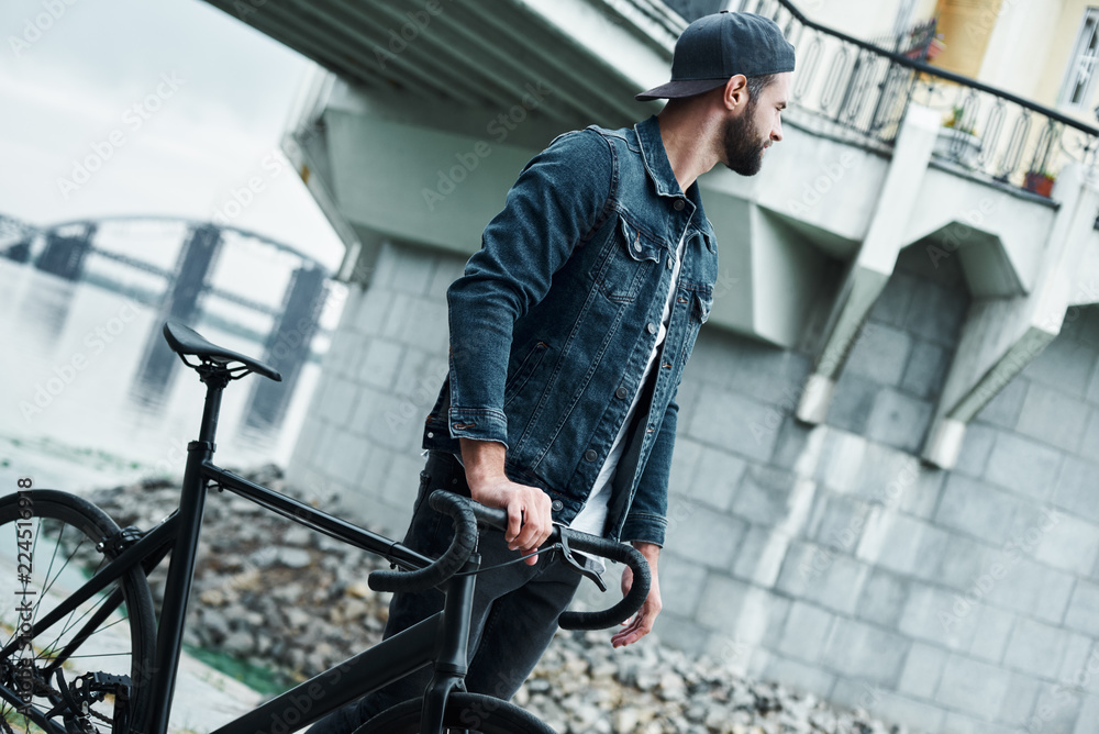 Outdoors leisure. Young stylish man walking on city street with bicycle looking aside pensive Outdoors leisure. Young stylish man walking on city street with bicycle looking aside pensive