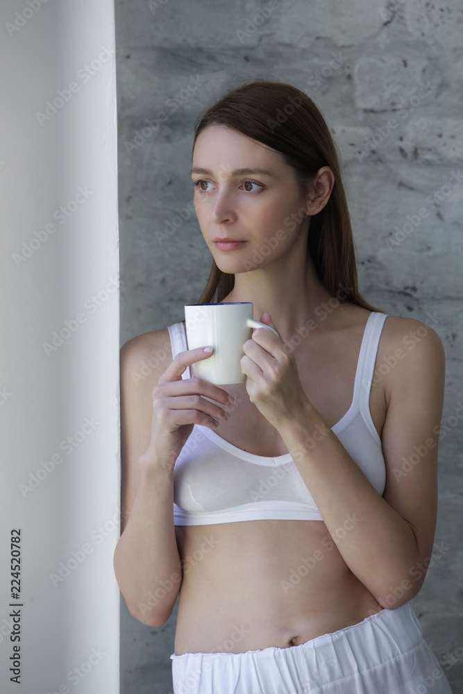 Young beautiful woman in casual cotton outfit leaning on the wall and drinking a tea or coffee, calm morning lifestyle concept Young beautiful woman in casual cotton outfit leaning on the wall and drinking a tea or coffee, calm morning lifestyle concept