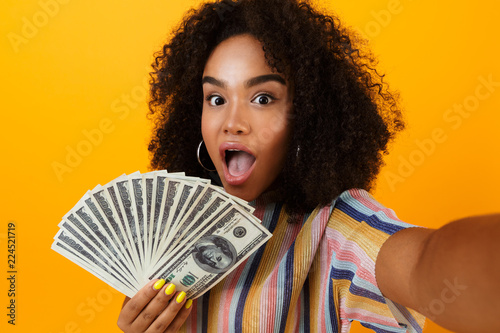 Excited young african woman posing isolated over yellow background holding money make selfie by camera. photo