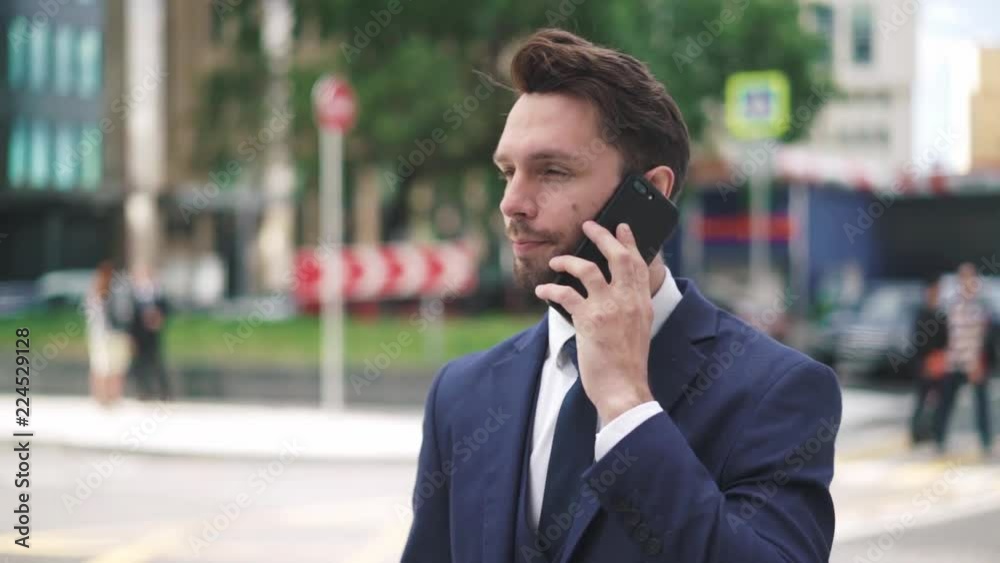 Hansome young caucasian businesman with fair hair and beard wearing a dark suit and tie talking on smartphone and smiling on a summer day. Hanheld real time medium shot Hansome young caucasian businesman with fair hair and beard wearing a dark suit and tie talking on smartphone and smiling on a summer day. Hanheld real time medium shot