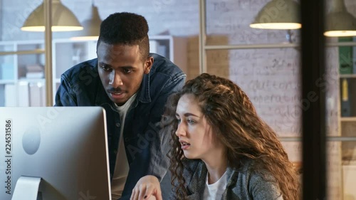 Tilt down of young pretty businesswoman pointing at computer screen and duscussing something with male photo