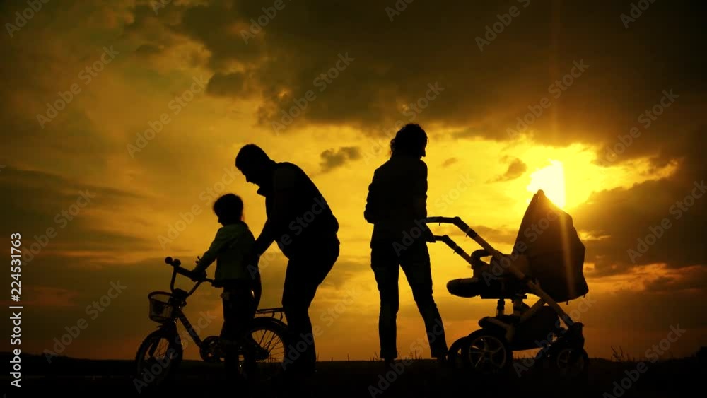 Silhouettes of happy parents having good time with their little children on the outdoor at sunset. Silhouettes of parents walk with small children with a stroller and bicycle.