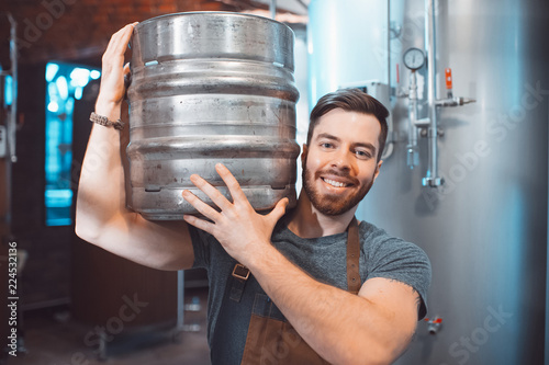Cuadro en lienzo A young brewer in an apron holds a barrel with beer in the hands of a brewery