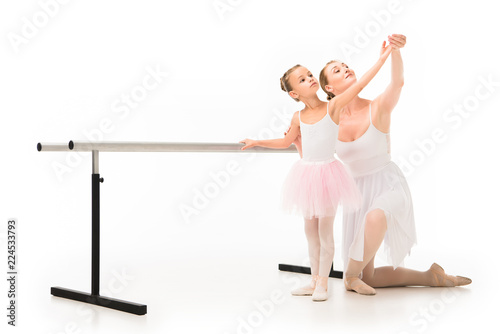 happy female teacher in tutu helping little ballerina exercising at ballet barre stand isolated on white background
