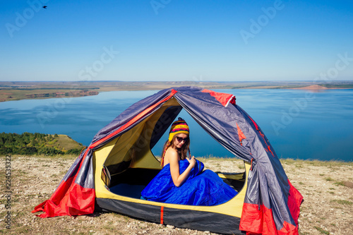 Back view of sexy naked woman tourist in a funny hat from Nepal sitting in blue sleeping bag in a tent sunny good morning in the mountains lake.