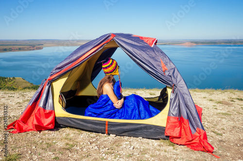 Back view of sexy naked woman tourist in a funny hat from Nepal sitting in blue sleeping bag in a tent sunny good morning in the mountains lake.