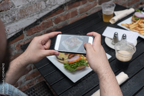 Point of view shot, man takes photo of food with mobile phone at an outdoor bar. Taking a picture of your food with your phone. Hamburger and fries on a white plate outside on a black table.