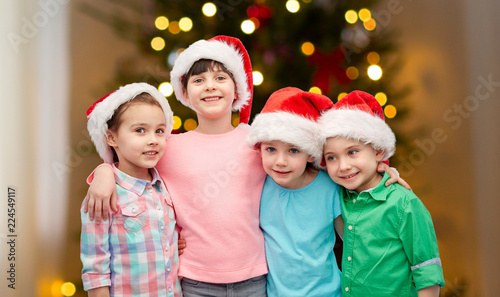 childhood, holidays, friendship and people concept - group of happy smiling little children in santa hats hugging over christmas tree lights background