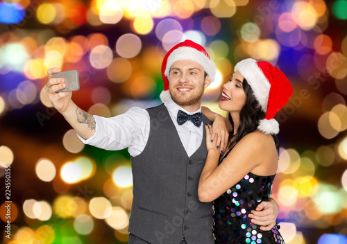 celebration, people and holidays concept - happy couple in santa hats taking selfie by smartphone at christmas or new year party over festive lights background