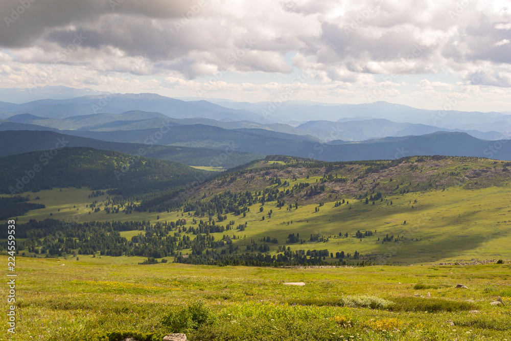 Landscape of green valley flooded with light with lush green grass, mountains, covered with stone and hills, a fresh summer day under a blue sky with white clouds and sun rays in Altai mountains