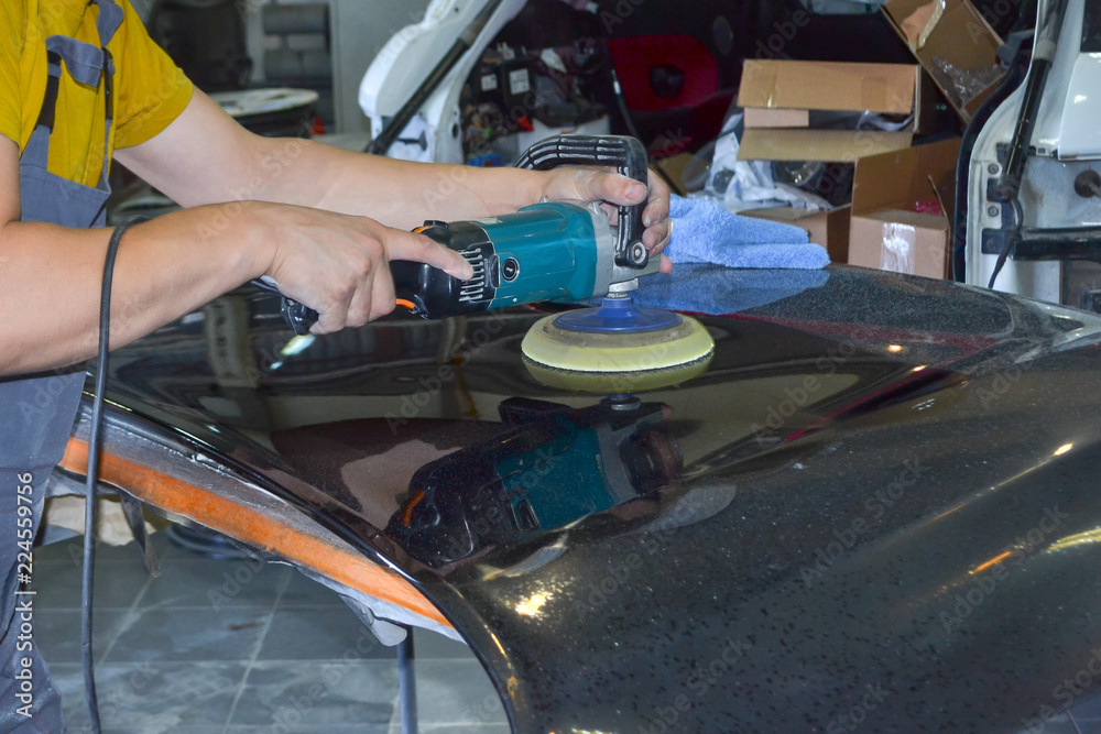 Fototapeta premium The master man of the detailing in work clothes and dirty hands polishes the bodywork of the bonnet of the car in black with a polishing machine in the workshop for body repair cars