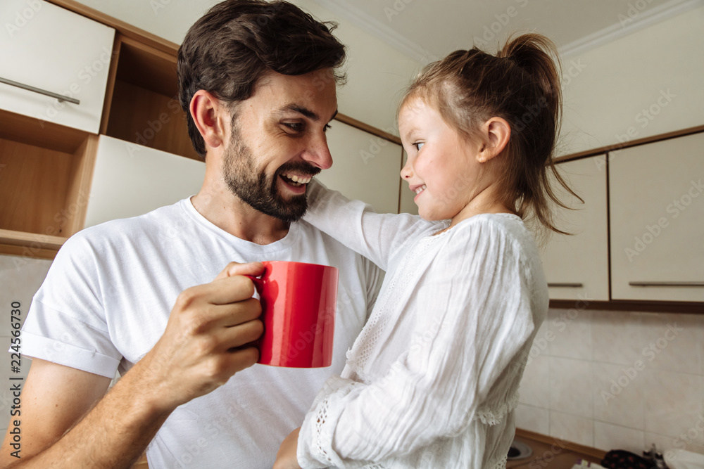 Parenthood. Family. Home. Dad is holding a cup and his little daughter in his arms. Both are talking and smiling. In the kitchen at home Parenthood. Family. Home. Dad is holding a cup and his little daughter in his arms. Both are talking and smiling. In the kitchen at home