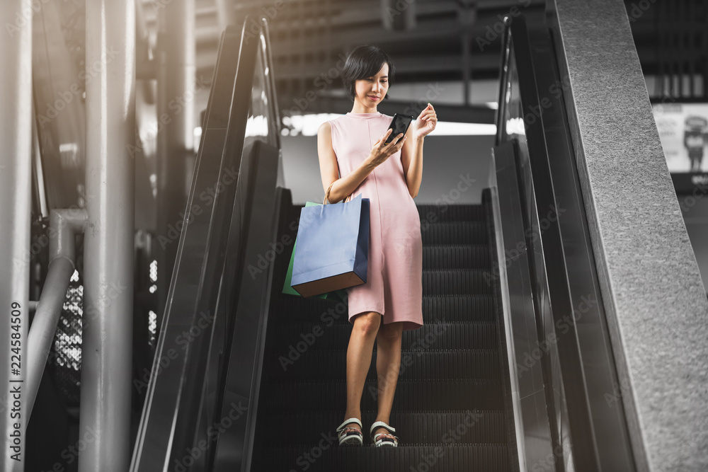 Young Asian businesswoman holding her shopping bags while using her smartphone for online shopping on the escalator in the shopping mall