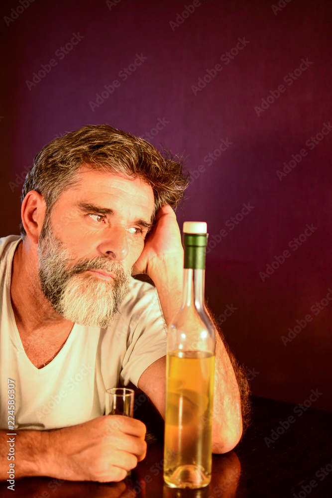 A depressed man with a beard sits at a table drunk and uncomfortable due to his addiction disease. Problems in marriage and at work. Alcohol addiction bad habits concept. Modern urban  lifestyle.