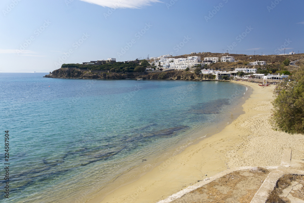 Colorful sandy beach with azure water and local white buildings. Agios Stefanos Beach of Mykonos island, Greece.