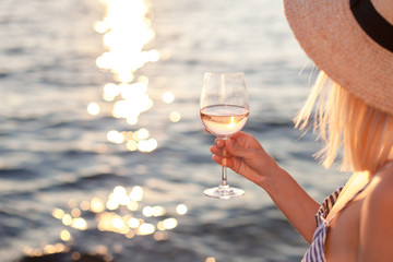 Girl holds wineglass with white wine in hand on beach at summer background of sunset sea or ocean. Blond beautiful woman is straw hat is relaxing, drinking, traveling and enjoying life in vacation.