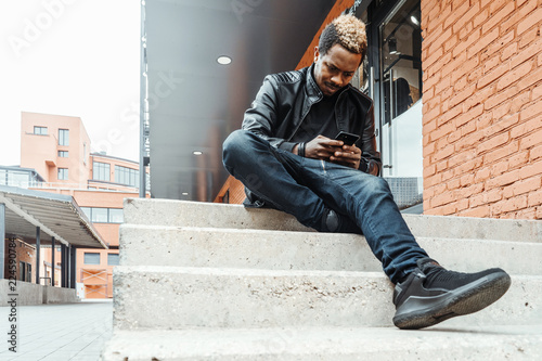 Dark-skinned smiling man looking at mobile phone screen and sitting on steps.
