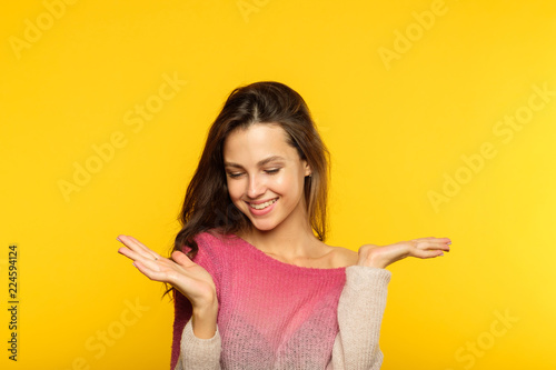 happy smiling joyful cute young woman holding virtual object on the palms of her hand. choice selection comparison concept. beautiful brown haired girl on yellow background with advertising space.