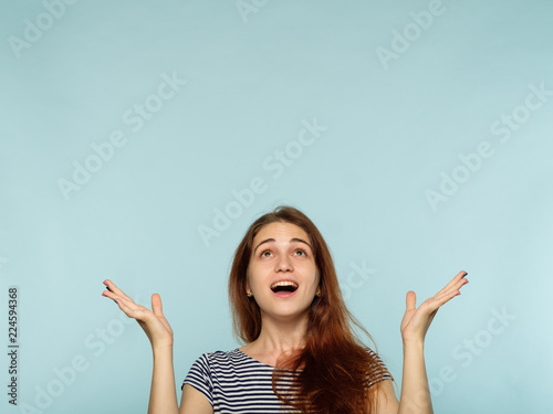 emotion face. happy joyful amazed woman with beaming smile. young beautiful brown haired girl pointing up above her head. advertising space on blue background.