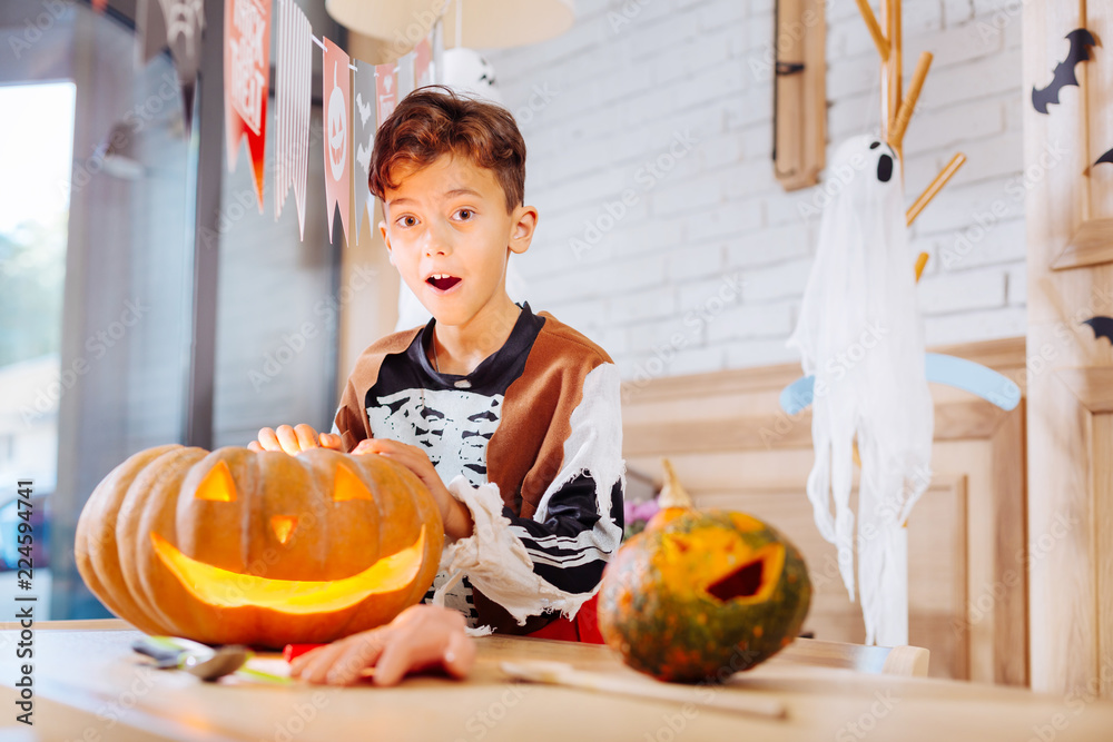 Fototapeta premium Excited boy. Cute funny boy wearing feeling skeleton costume excited while standing near carved Halloween pumpkin