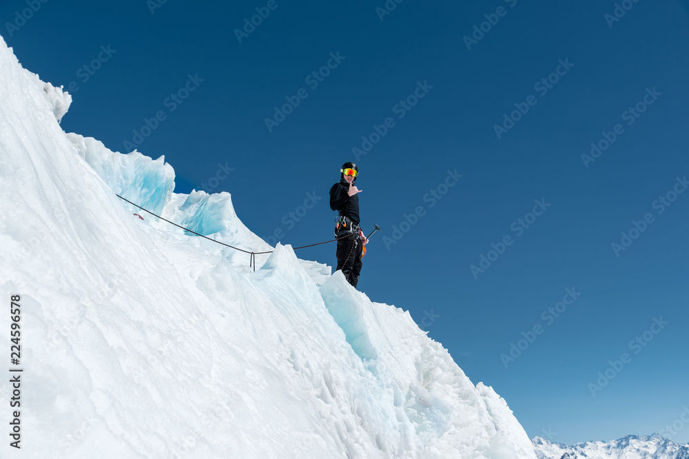 A mountaineer stands on the edge of a glacier with a snow shovel in his hands and shows Shak's gesture against the blue sky