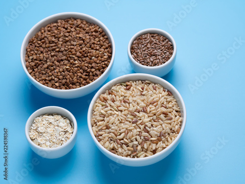 brown rice, buckwheat, flax and oat flakes in white cups on a blue background