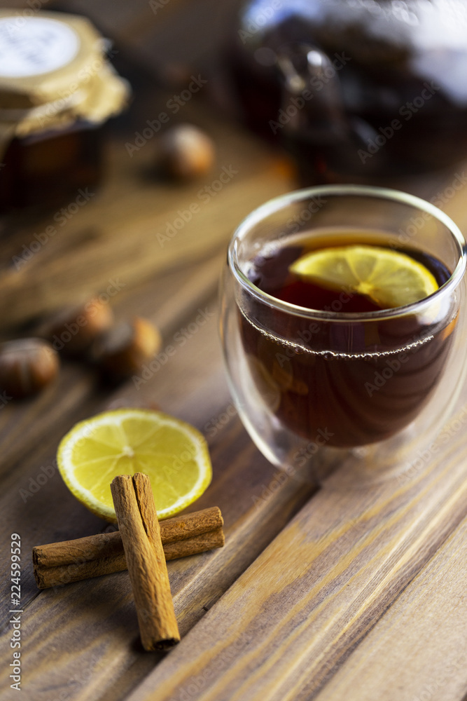 Tea in glass cup with lemon and vanilla pod at wood background. teapot with tea near. Huzelnut and jam in jar at wood board behind. Tea in glass cup with lemon and vanilla pod at wood background. teapot with tea near. Huzelnut and jam in jar at wood board behind.