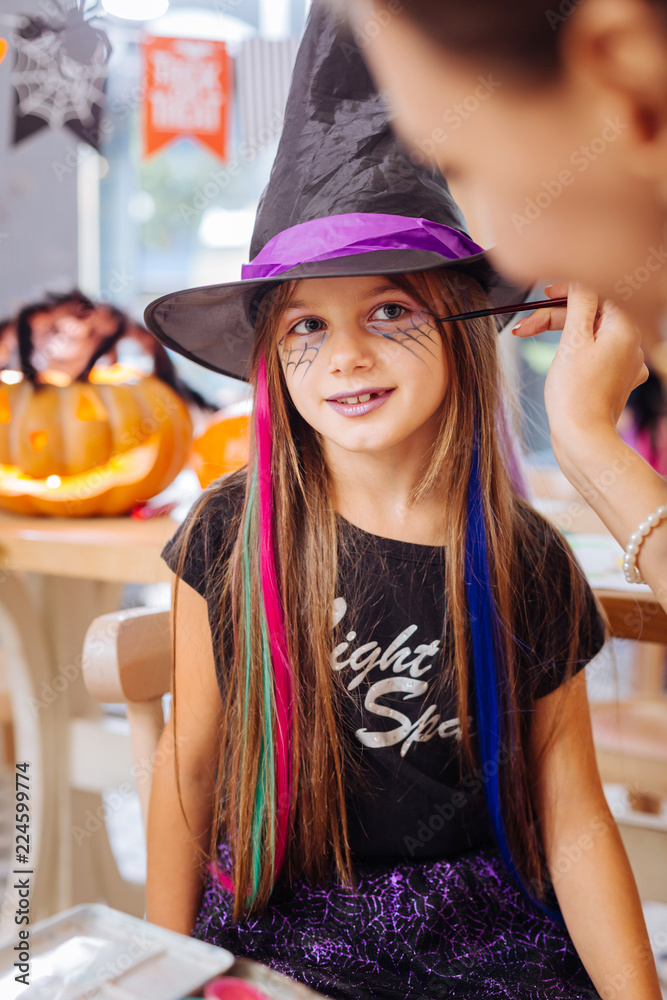Halloween makeup. Dark-haired beautiful cute girl wearing wizard hat feeling happy getting Halloween makeup