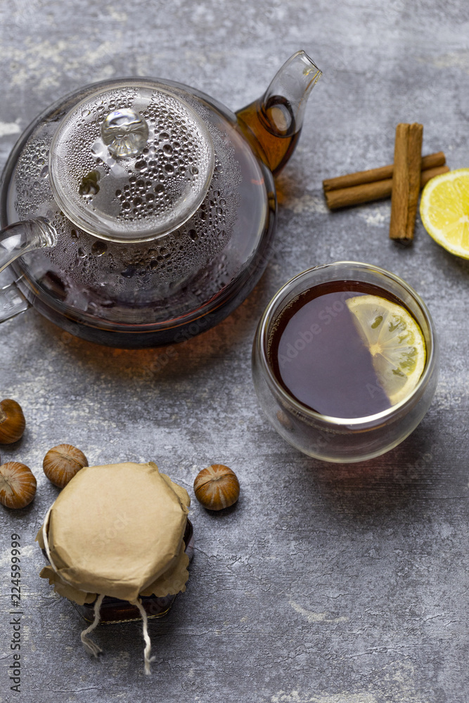 Tea in glass cup with lemon and vanilla pod at wood background. teapot with tea near. Huzelnut and jam in jar at wood board behind. Tea in glass cup with lemon and vanilla pod at wood background. teapot with tea near. Huzelnut and jam in jar at wood board behind.