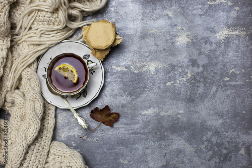 Cup with black tea and lemon and saucer, spoon, jelly in jar, maple leaves and knitted scarf near, an gray background