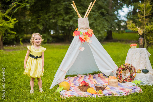 Little girl lying and playing in a tent, children's house wigwam in park Autumn portrait of cute curly girl. Harvest or Thanksgiving. autumn decor, party, picnic. Child in yellow dress with apple 