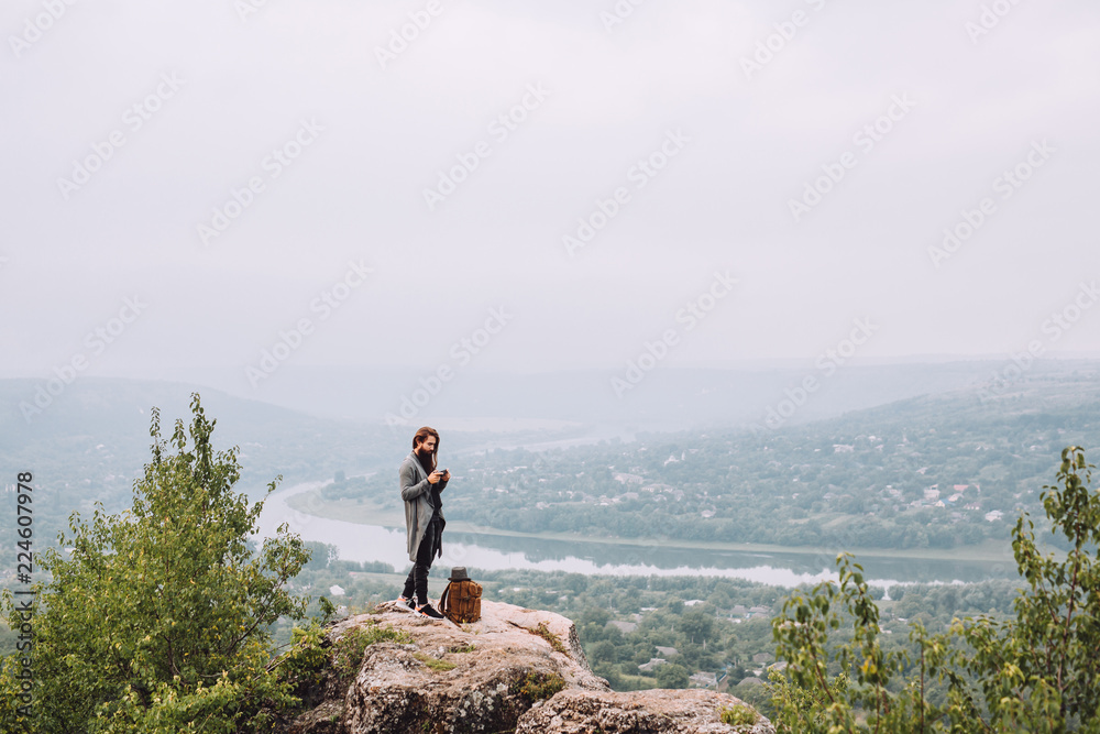 Young tourist with a beard looks at the pictures that he made on a digital camera.Man dressed stylish clothes in the background beautiful summer landscape