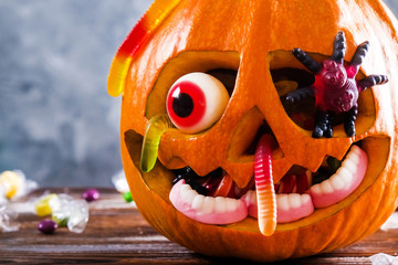 Orange pumpkin carved into Jack o Lantern with eyeball candy and gummy worms & spiders on wooden table. Background, copy space, close up, top view. Halloween party decoration. Trick or treat concept.