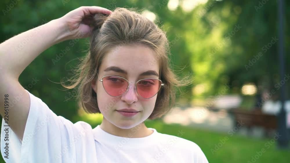 Attractive young woman in pink sunglasses and white t shirt taking off her rubber band and playing with her hair in summer park. Concept of fashion and accessories. Slider slow motion portrait shot Attractive young woman in pink sunglasses and white t shirt taking off her rubber band and playing with her hair in summer park. Concept of fashion and accessories. Slider slow motion portrait shot