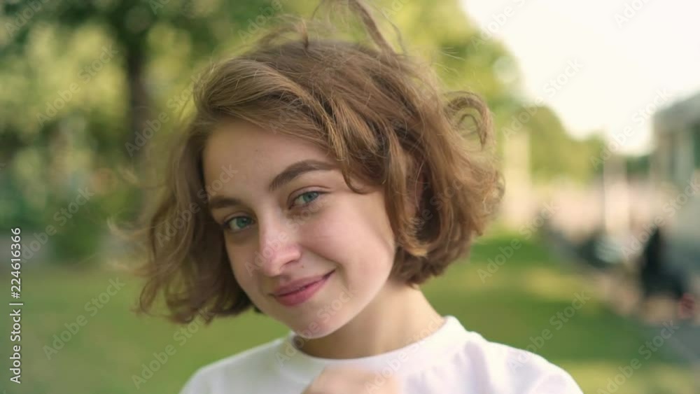 Attractive young girl with short wavy hair smiling and winking looking at camera standing in summer park. Flirting and seduction concept. Right to left pan slow motion close up shot