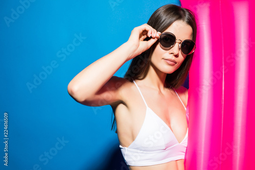 Beautiful young smiling woman dressed in white swimsuit and glasses posing with pink inflatable mattress isolated over blue background